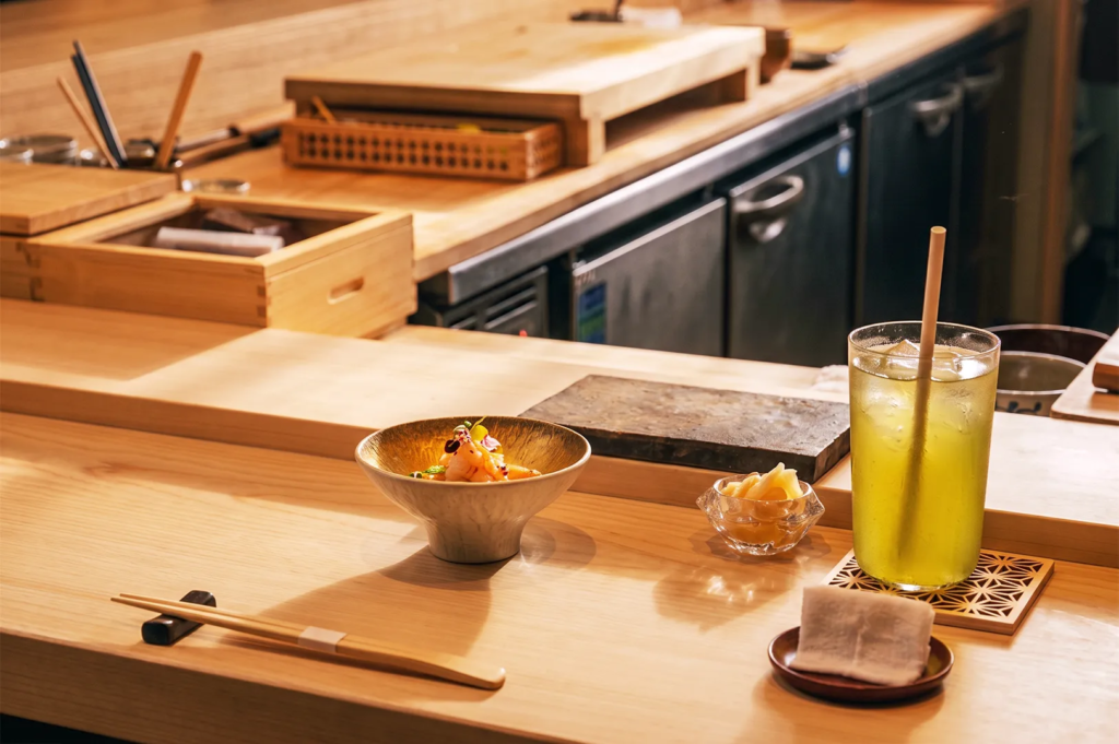 Minimalist dining setup featuring a golden bowl of seafood appetizer, iced green tea, pickled ginger, and chopsticks on a light wooden counter in a serene omakase restaurant.