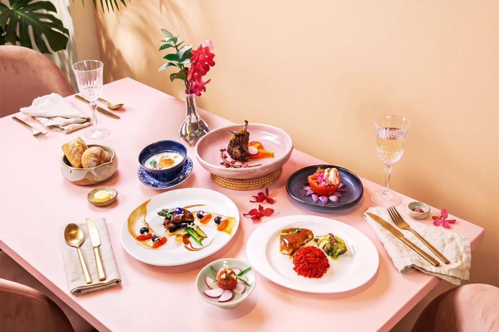 Stylish pink table setup with artfully plated modern cuisine, gold cutlery, crystal glasses, and floral decor, emphasizing creative plating and luxury dining aesthetics.