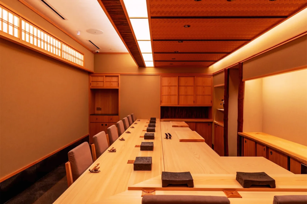 Private omakase dining room in Singapore with long wooden counter, cushioned chairs, and minimalist Japanese interior design featuring warm lighting and natural wood tones.