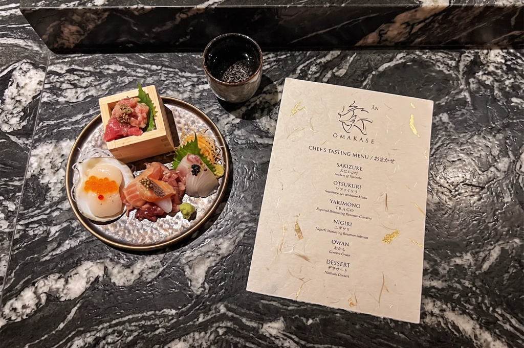 Artistic plate of assorted sashimi with salmon roe and tuna tartare beside a printed omakase chef’s tasting menu on a marble countertop, showcasing fine Japanese dining elegance.