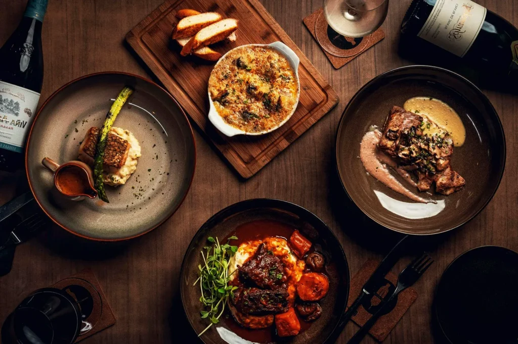 Gourmet fine dining spread in Singapore featuring seared fish, braised beef, gratin, and wine pairings on a wooden table under warm lighting.