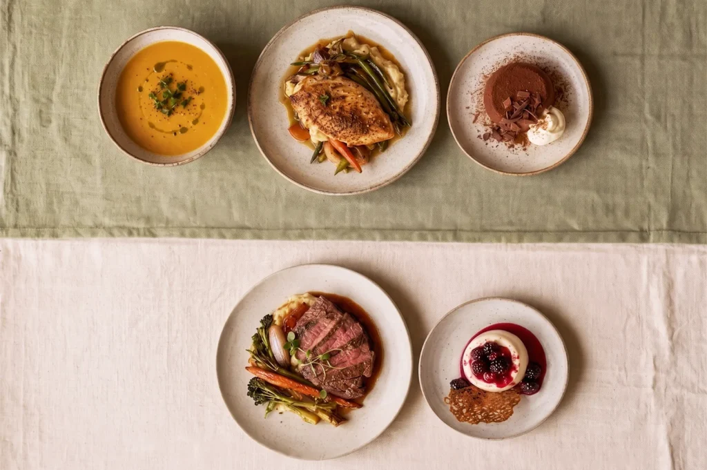 Overhead view of a balanced fine dining menu including creamy soup, roasted chicken, beef steak with vegetables, and chocolate and berry desserts, highlighting modern European cuisine.