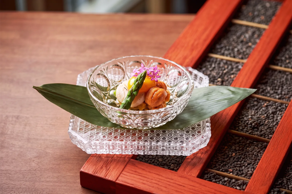 Delicate Japanese dish with sea urchin, scallops, asparagus, and edible flowers in glass bowl — elegant fine dining plating.